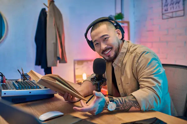 Un uomo asiatico sorridente che indossa podcast per cuffie nel suo studio di casa, con un microfono e un mixer davanti a lui. — Foto stock