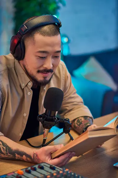 A handsome Asian man wearing headphones and a shirt reads a book while recording a podcast in his studio. — Stock Photo