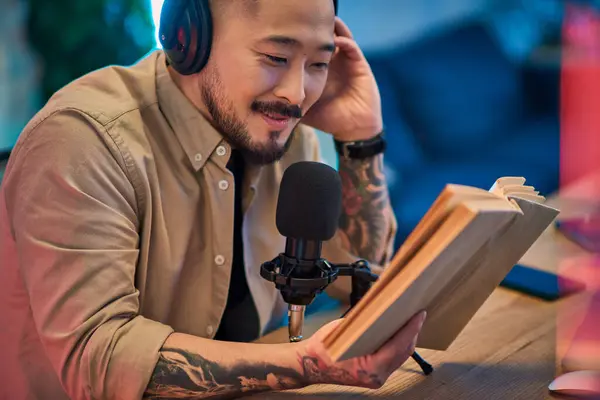 An Asian man sits at a desk in his home studio, wearing headphones and holding a book while recording a podcast. — Stock Photo