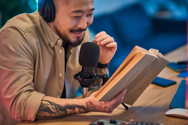 A handsome Asian man sits in his podcast studio, reading from a book while recording. — Stock Photo