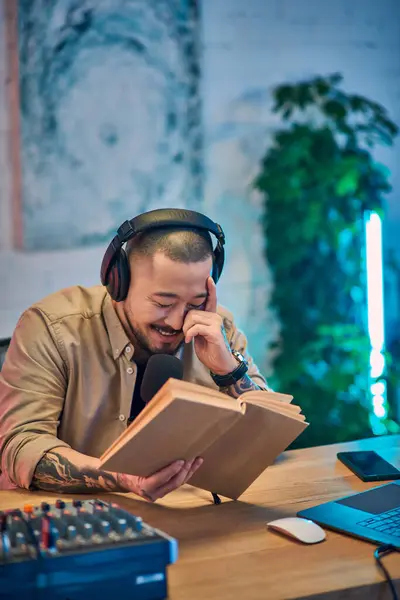 An Asian man with headphones smiles while reading from a book in his podcast studio. — Stock Photo