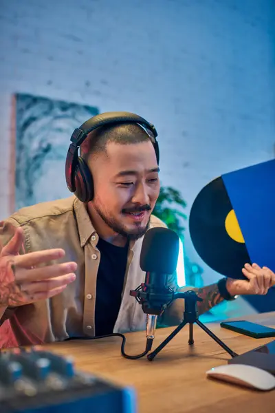 A young Asian man wearing headphones and a beige shirt is recording a podcast in his studio. He is speaking into a microphone and gesturing with his hands. — Stock Photo