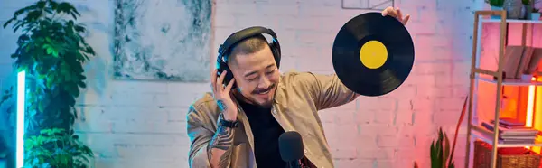 A handsome Asian man podcasting in a brightly lit studio, holding a vinyl record. — Stock Photo