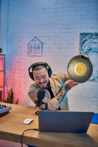 Un giovane asiatico sorride e regge un disco in vinile mentre registra un podcast nel suo studio. — Foto stock