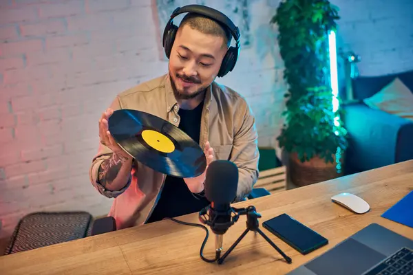 A handsome Asian man wearing headphones holds a vinyl record in his studio while recording a podcast. — Stock Photo