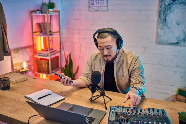 A handsome Asian man records a podcast in his home studio, adjusting the soundboard with his hand. — Stock Photo