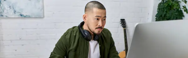 A handsome Asian man, wearing headphones, focuses intently on a computer screen in his studio, surrounded by instruments. — Stock Photo