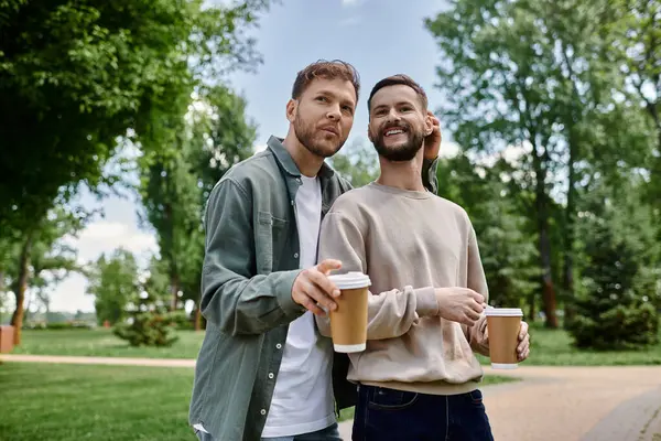 Dois barbudo gay homens no casual traje stand no um parque, segurando café copos e sorrindo. — Fotografia de Stock