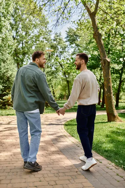 A bearded gay couple walks hand-in-hand on a paved path in a green park, laughing and enjoying a sunny day together. — Stock Photo