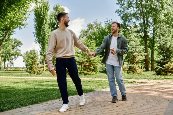 Two bearded gay men holding hands and smiling while walking through a green park, enjoying coffee on a sunny day. — Stock Photo
