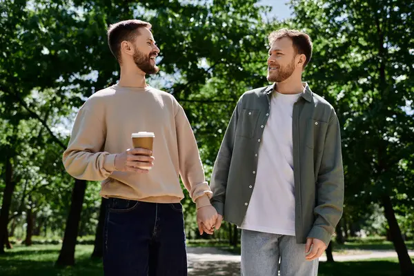 Deux hommes barbus, vêtus avec désinvolture, marchent main dans la main dans un parc verdoyant tout en dégustant un café. — Photo de stock