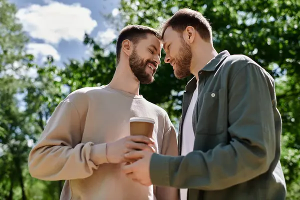 Dois homens barbudos compartilham um café e um momento amoroso em um parque verde em um dia ensolarado. — Stock Photo