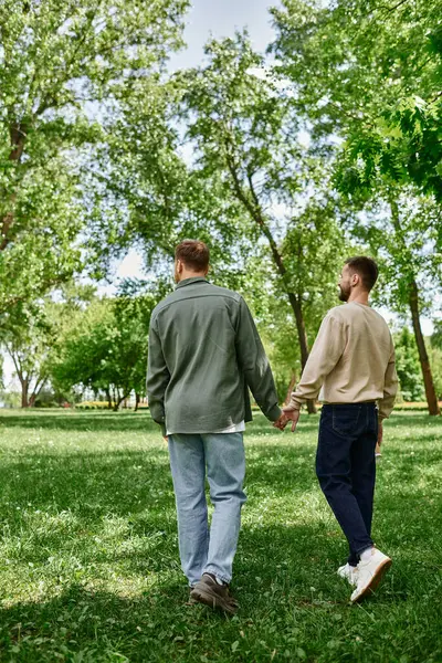 A gay couple with beards, dressed in casual attire, walks hand-in-hand through a verdant park. — Stock Photo