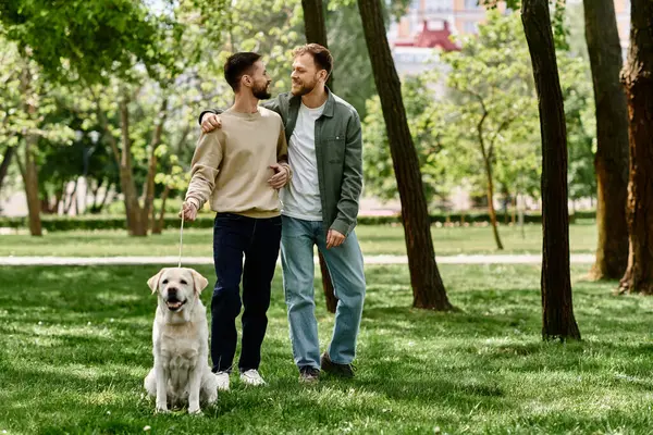 A bearded gay couple walks hand-in-hand through a green park with their labrador retriever. — Stock Photo