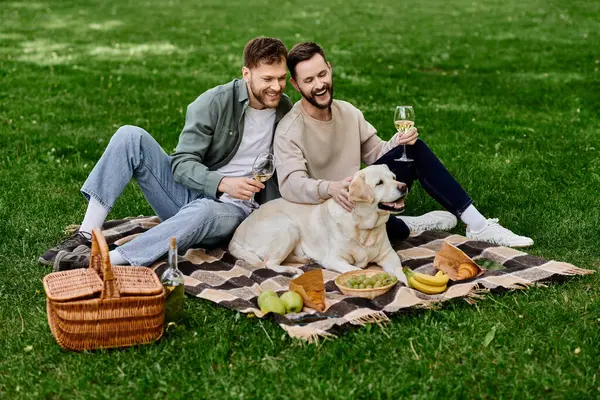 A bearded gay couple enjoys a picnic in a green park with their Labrador Retriever. They are sitting on a blanket and drinking wine while their dog rests beside them. — Stock Photo
