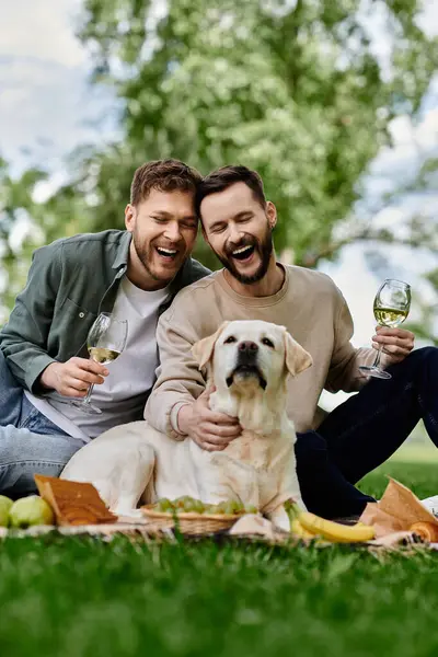 Um casal gay barbudo desfruta de um piquenique com seu Labrador retriever em um exuberante parque verde, compartilhando risos e um copo de vinho. — Fotografia de Stock