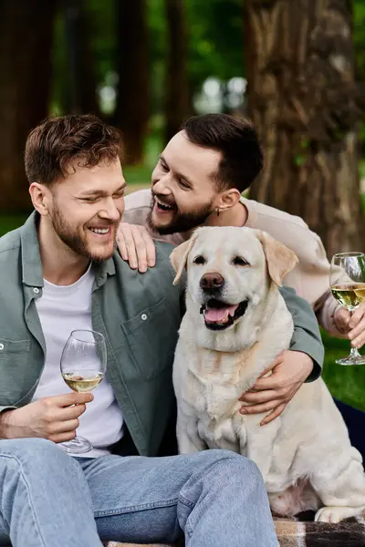 A bearded gay couple laughs with their labrador dog while enjoying a picnic in a green park. — Stock Photo