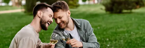 A bearded gay couple enjoys a picnic in a lush green park while sharing a laugh and a glass of wine. — Stock Photo