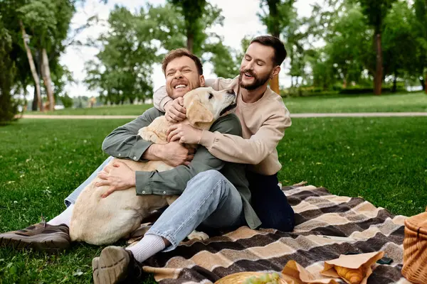 A happy bearded gay couple enjoys a picnic with their labrador dog in a green park. — Stock Photo
