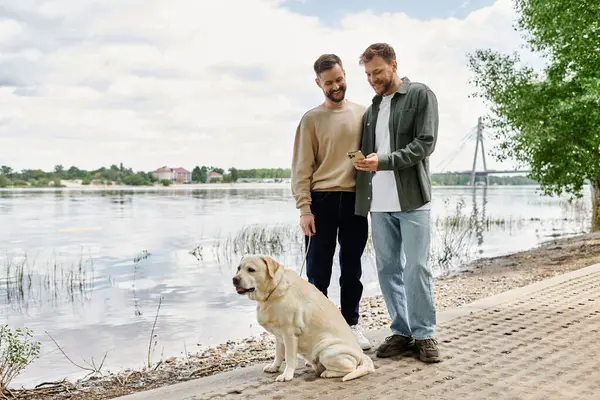 A bearded gay couple enjoys a walk with their labrador dog by a lake on a sunny day. — Stock Photo
