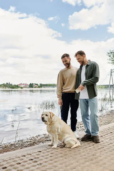 A bearded gay couple enjoys a walk with their labrador dog by a lake on a sunny day. — Stock Photo