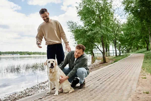 Zwei bärtige Männer genießen einen Spaziergang in einem grünen Park mit ihrem Labrador Retriever am Wasser. — Stockfoto