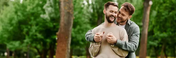 Una coppia gay barbuta si abbraccia felicemente in un lussureggiante parco verde, circondato da alberi. — Foto stock