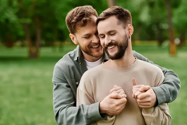 Due uomini barbuti, una coppia gay, si tengono per mano e ridono in un lussureggiante parco verde. — Foto stock