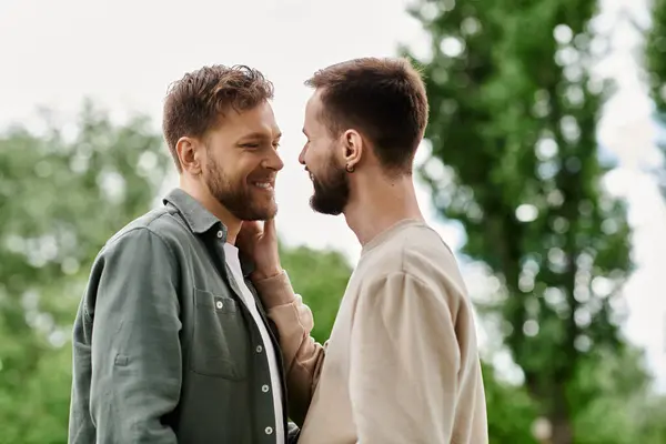 Due uomini barbuti condividono un momento d'amore in un lussureggiante parco verde. Sono vicini, guardandosi negli occhi con affetto. — Foto stock