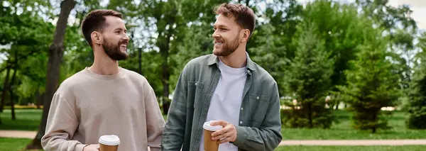 Two bearded men, dressed casually, walk together in a park, smiling and holding coffee cups. — Stock Photo