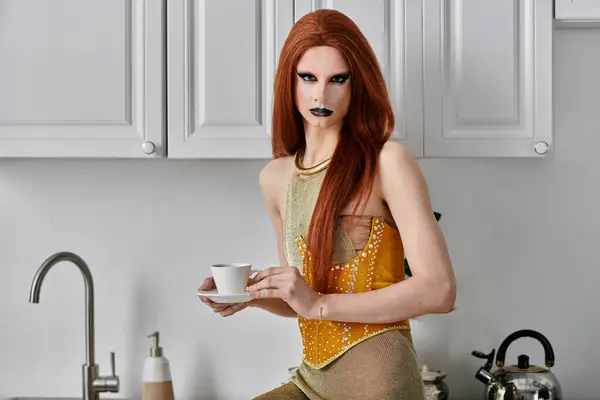 A glamorous drag diva in a yellow corset and long red hair holds a cup of coffee in a modern kitchen. — Stock Photo