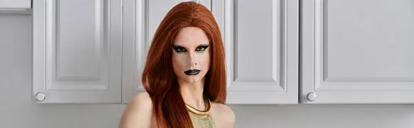 A drag queen poses in a kitchen with white cabinets, wearing a glamorous outfit. — Stock Photo