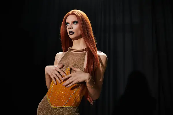 Una drag queen glamour in un abbagliante vestito d'oro e arancione si erge su uno sfondo scuro. — Foto stock