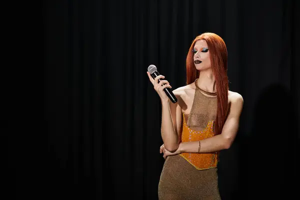 Un drag performer con lunghi capelli rossi e un abito glamour tiene un microfono davanti a una tenda nera. — Foto stock