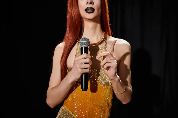 A drag queen in a gold sequin dress holds a microphone and points upwards. — Stock Photo
