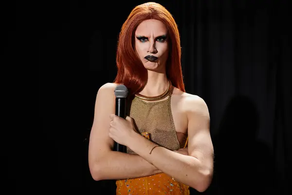 A drag performer with red hair and striking makeup holds a microphone, posing confidently on a dark stage. — Stock Photo