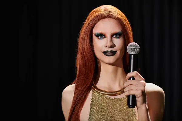Una drag queen con pelo rojo largo y maquillaje audaz sostiene un micrófono antes de una actuación. - foto de stock