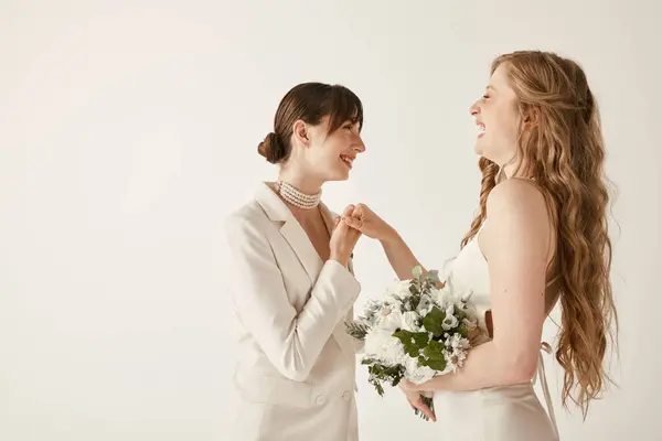 Duas noivas de vestido de casamento branco de pé de mãos dadas e sorrindo um para o outro. — Fotografia de Stock