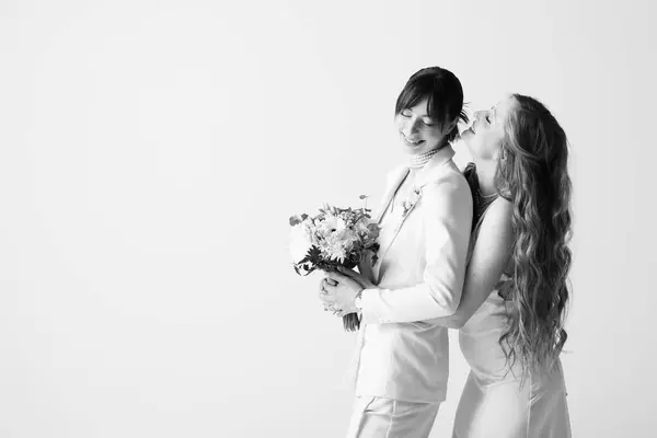 Two brides, dressed in white, embrace on their wedding day. The bride on the left holds a bouquet of flowers and smiles at her partner. — Stock Photo