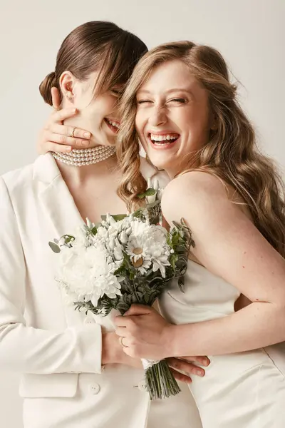 Duas noivas de traje branco abraçam e riem, segurando um buquê de flores brancas. — Fotografia de Stock