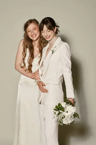 Dos mujeres vestidas de blanco traje de novia, sonrientes y abrazadas, posan sobre un fondo gris. - foto de stock