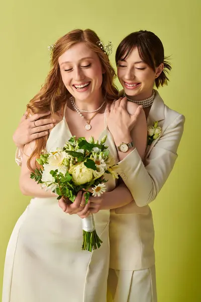 Two women in white attire embrace on their wedding day. The bride holds a bouquet of flowers while they smile at each other in front of a green background. — Stock Photo