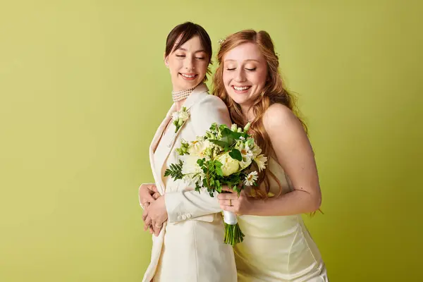 Two women dressed in white wedding attire pose against a green background. One woman holds a bouquet of flowers. — Stock Photo