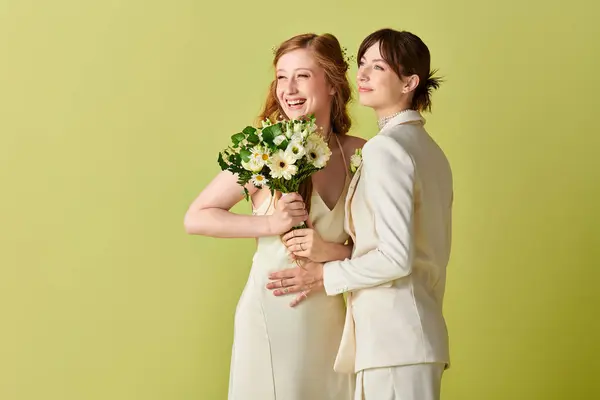 Two women in white wedding attire embrace, laughing with joy on a green backdrop. One holds a bouquet of white flowers. — Stock Photo