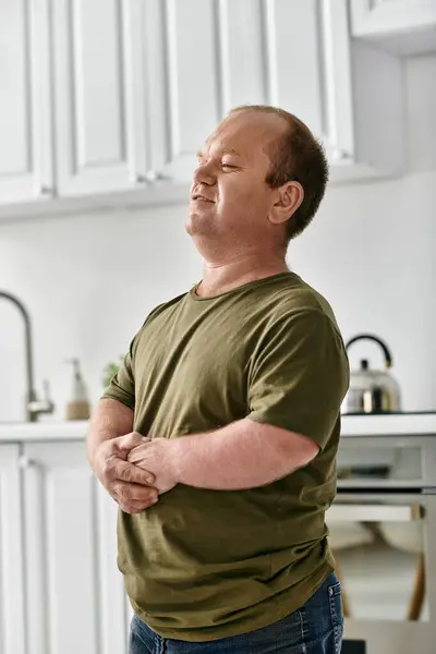 Un hombre con inclusividad se para en una cocina, vistiendo atuendo casual, con los ojos cerrados y las manos cerradas. - foto de stock