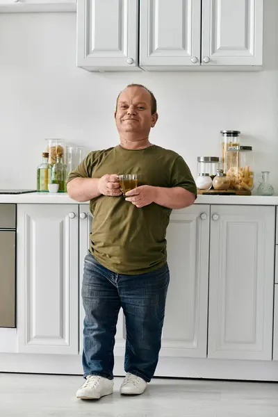 Чоловік з інклюзивністю стоїть на своїй кухні, тримаючи келих напоїв і посміхаючись. — стокове фото