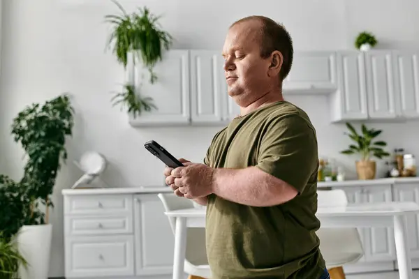 Un homme avec inclusivité se tient dans sa cuisine, vêtu avec désinvolture, tenant son téléphone et regardant vers le bas l'écran. — Photo de stock