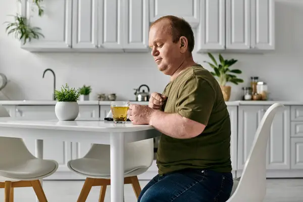 A man with inclusivity sits at a kitchen table, casually dressed, enjoying a cup of tea. — Stock Photo