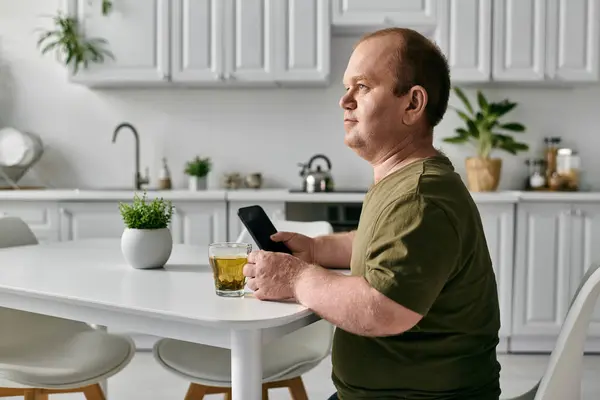 A man with inclusivity sits at a table in his kitchen, enjoying a cup of tea while looking at his phone. — Stock Photo
