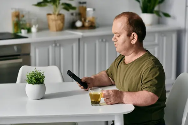 A man with inclusivity in casual clothing sits at a table in his home, enjoying a cup of tea while looking at his phone. — Stock Photo
